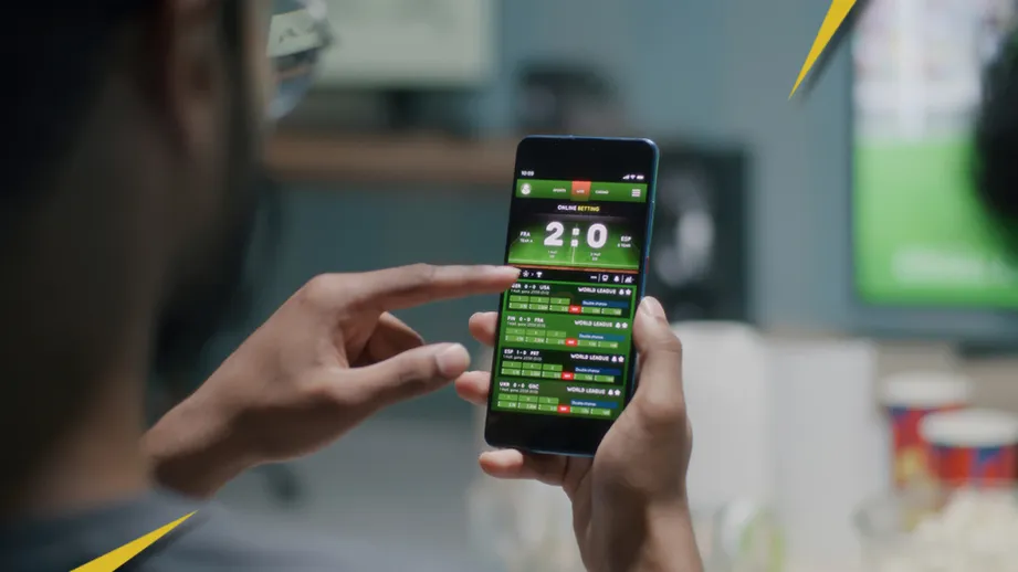 Melhores Apps de Apostas em Futebol, homem segurando um celular com uma aposta e a imagem é bem colorida.