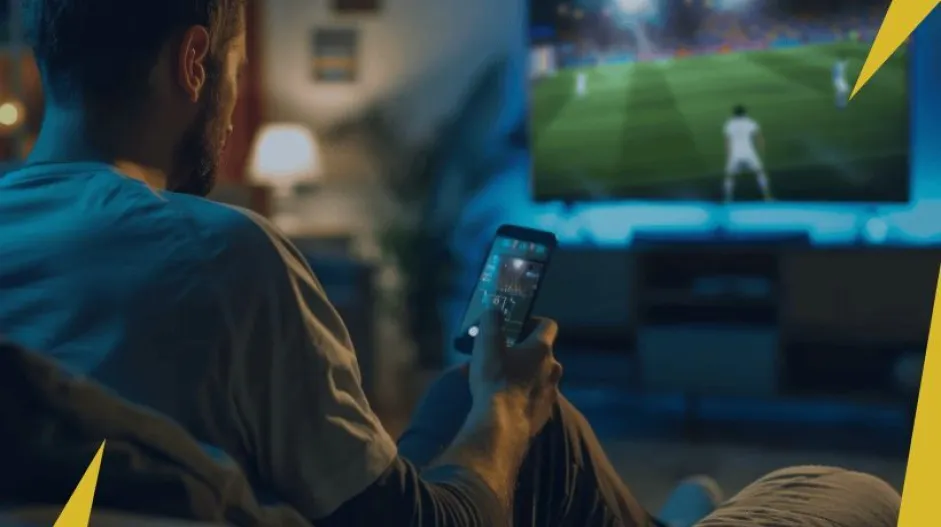 Google Pay como funciona: homem assistindo um jogo de futebol e com o celular em sua mão direita