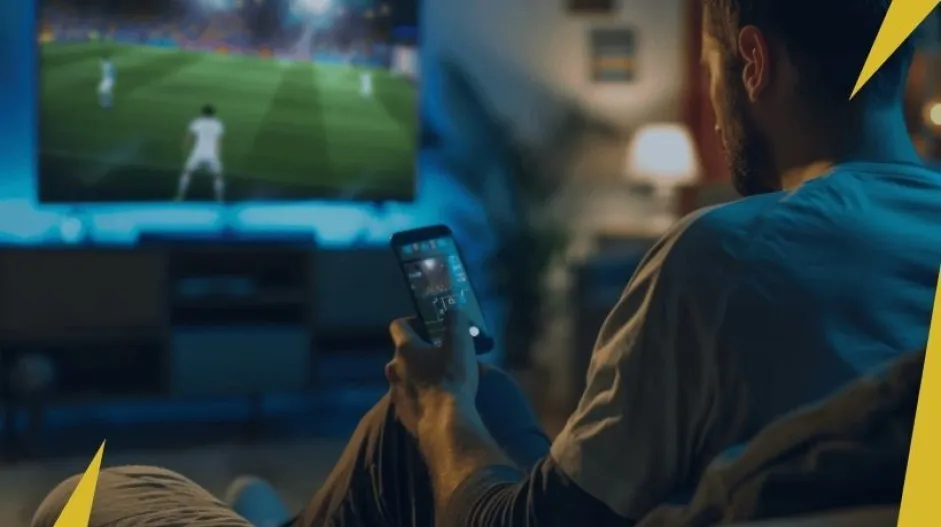 Uma mão segurando um celular em frente a uma televisão transmitindo futebol, com um abajur ligado ao fundo